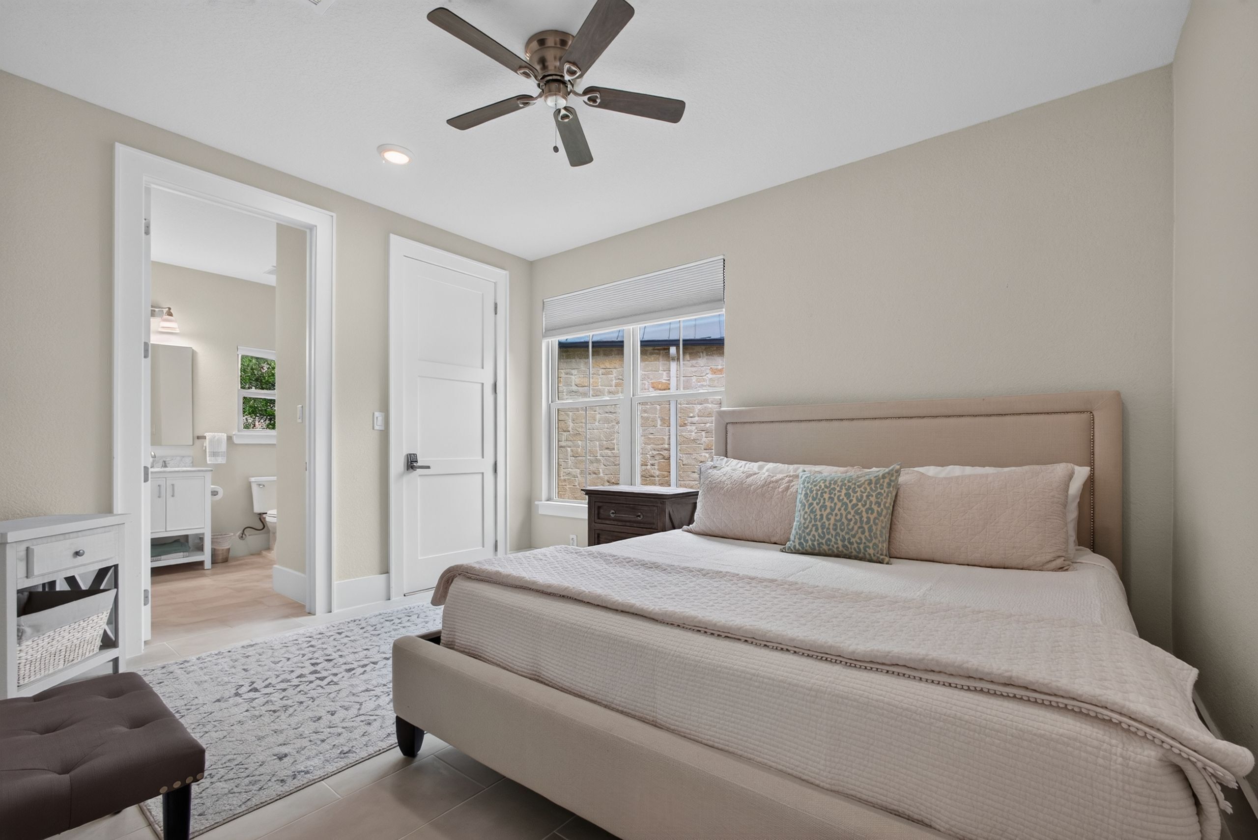 A bedroom with a bed, a ceiling fan, a window with a view, and a bathroom visible through an open door.