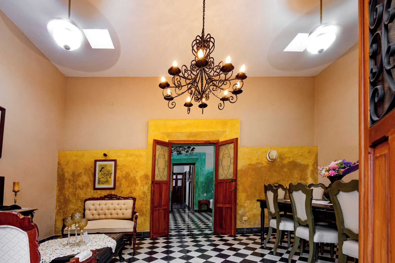A hotel room with a chandelier, a dining area with chairs, a checkered floor, and a view through an open door.