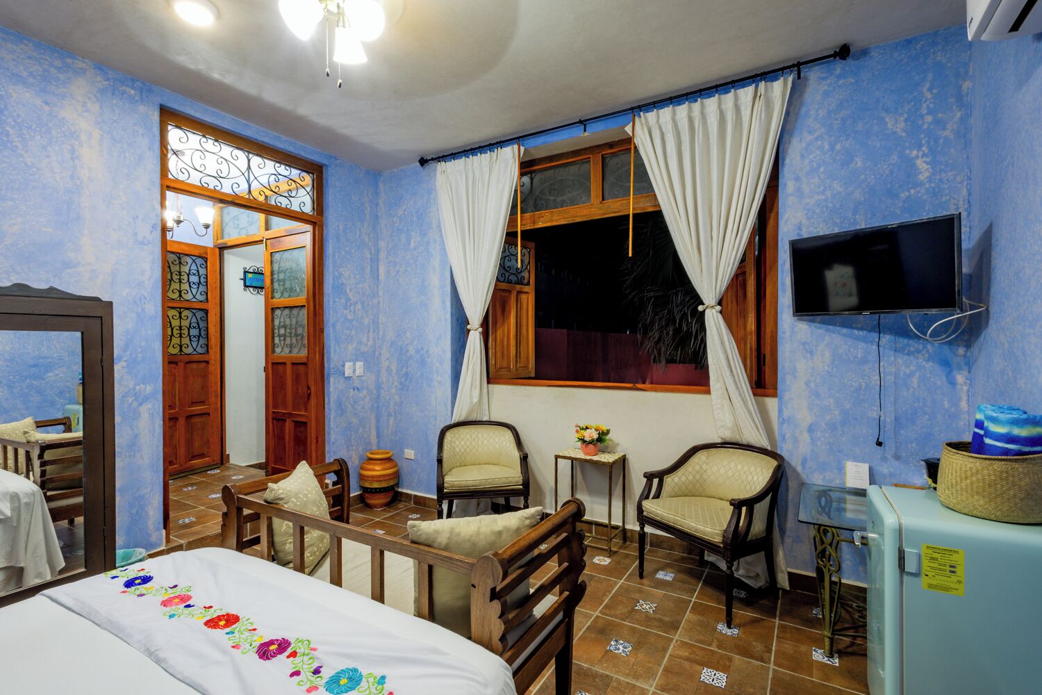 A room with a bed, two chairs, a small table with a flower vase, a television mounted on the wall, and a window with curtains.