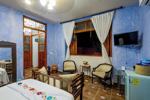 A room with a bed, two chairs, a small table with a flower vase, a television mounted on the wall, and a window with curtains. - Casa Vicenta (Izamal)