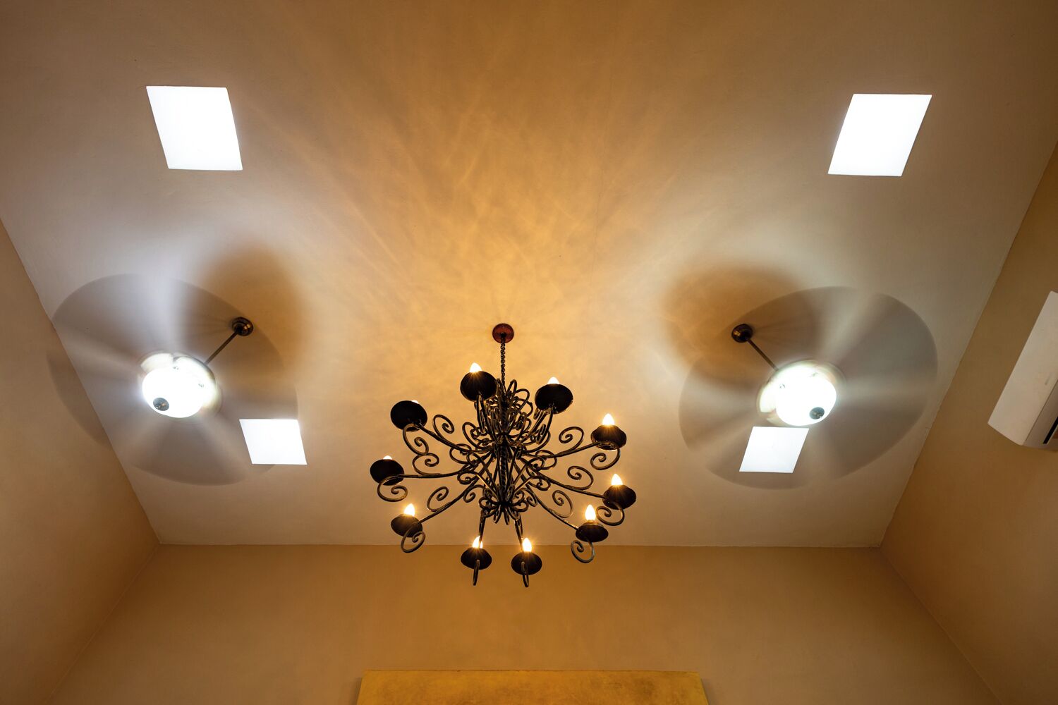 A room with a central chandelier, recessed ceiling lights, and a plain wall.