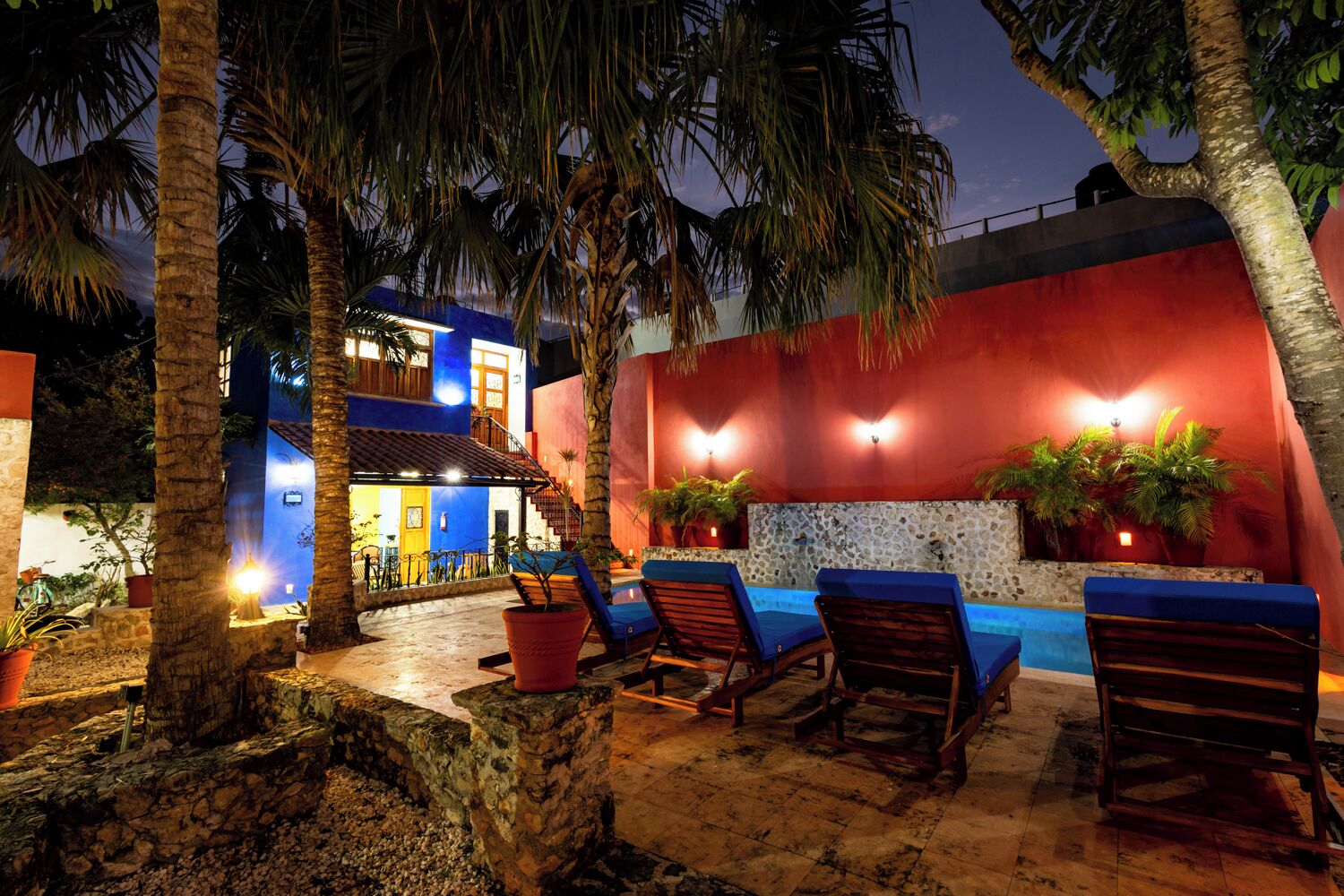 Zona junto a la piscina con sillones, piscina y edificio con pared roja y detalles azules.