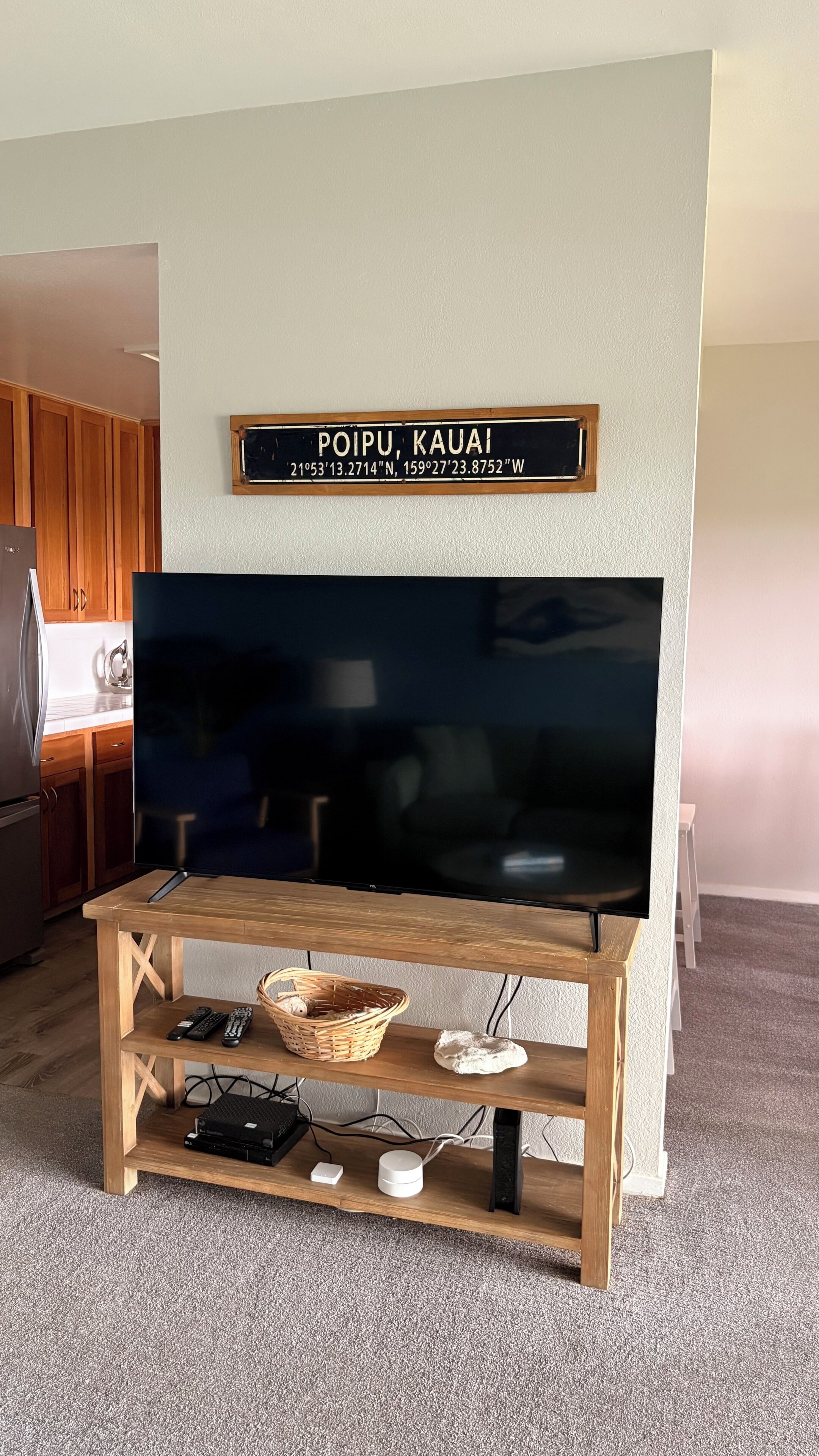 Un salon avec un téléviseur à écran plat posé sur un meuble télé en bois, une cuisine en arrière-plan et une enseigne murale indiquant l’emplacement : « POIPU, KAUAI ».