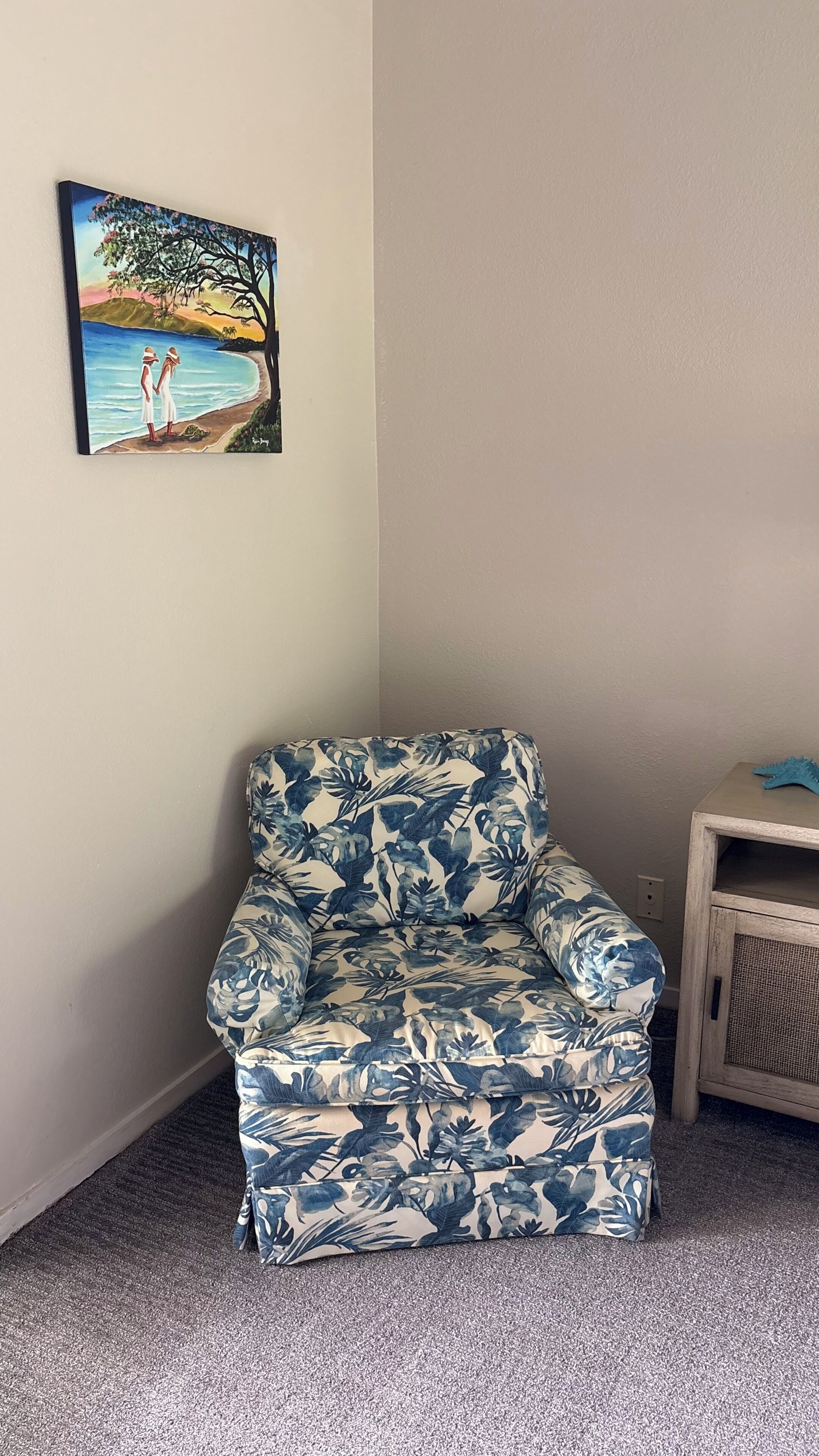 Een kamer met een fauteuil met bloemenprint, een schilderij van een strandgezicht en een klein houten kastje.