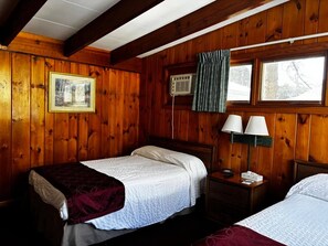 A wooden cabin-style room with two beds, a framed picture on the wall, an air conditioning unit, and a window with curtains.