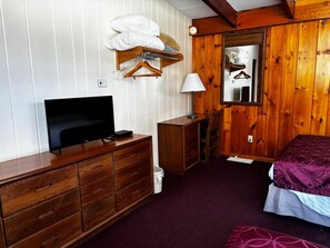 A room with a wooden dresser, a television, a bed with a patterned bedspread, a mirror, and a hanging clothes rack.