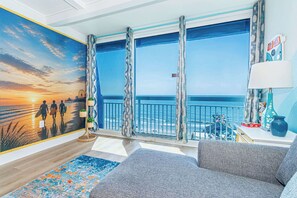 A room with a large window, a sofa, a small table, a lamp, and a mural of a beach scene. - Penthouse on 14th Ave Pier! 1BR Yachtsman 1101 (Myrtle Beach)