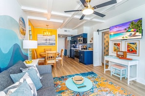 A modern living room with a sofa, a dining area, a kitchen, and a TV displaying a beach scene. - Penthouse on 14th Ave Pier! 1BR Yachtsman 1101 (Myrtle Beach)