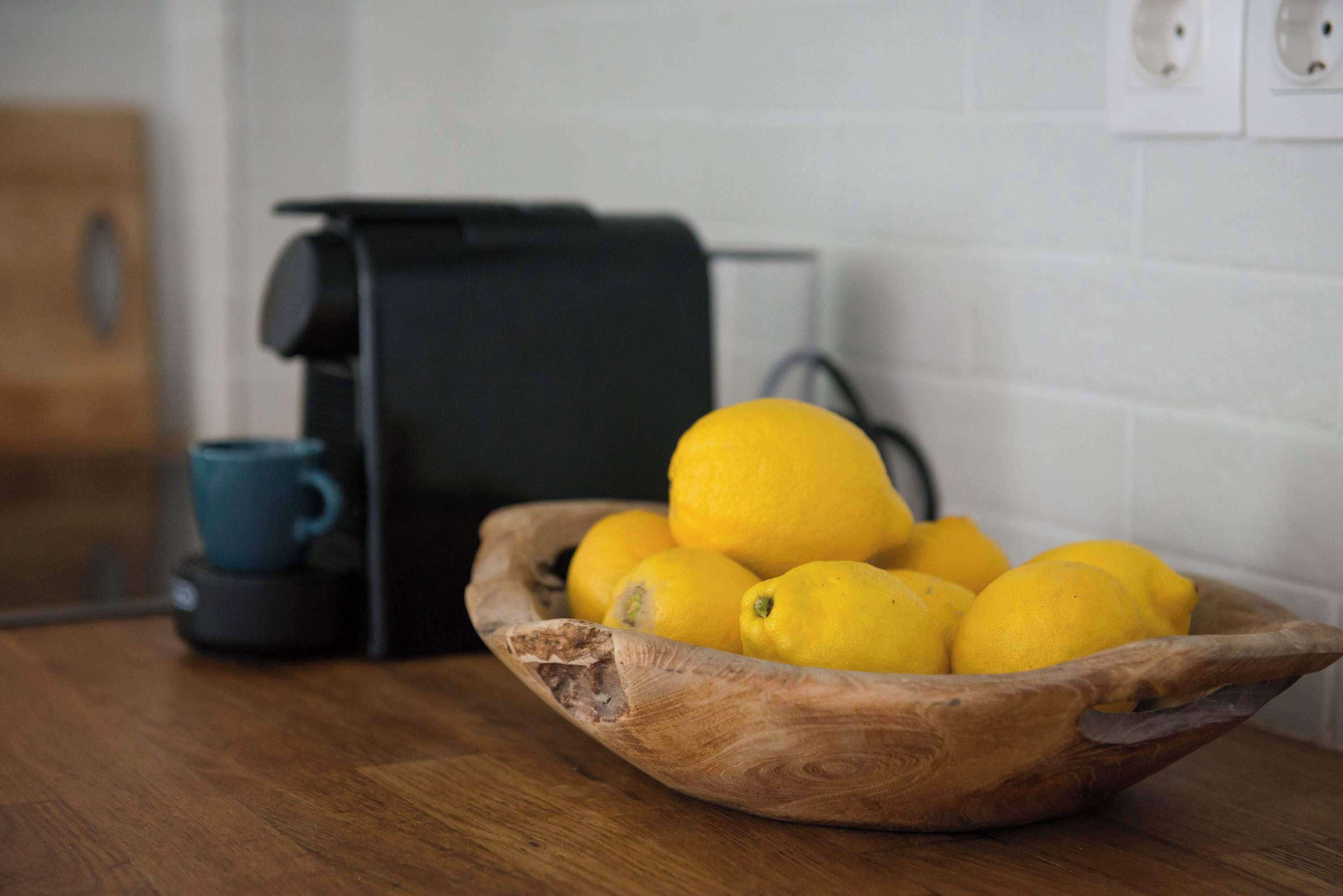 A wooden bowl with lemons on a kitchen counter, a blue mug, and a black appliance.