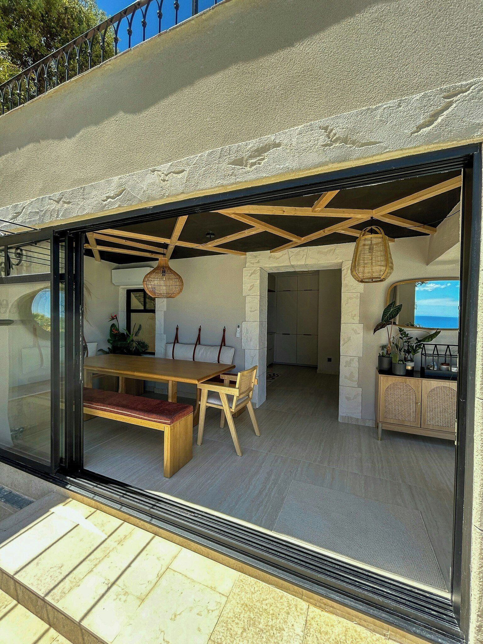 A modern interior with a wooden dining table, chairs, and a bench, a kitchen area with cabinets, and a view through a glass door.