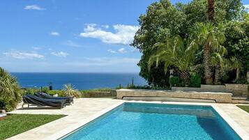 Une piscine rectangulaire à l’eau d’un bleu cristallin, entourée d’une terrasse dallée, de palmiers et offrant une vue sur l’océan.