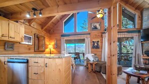 Interior de una cabaña de madera con cocina, comedor y un ventanal grande que da a un paisaje.