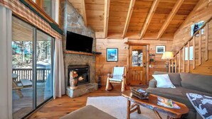 Interior de una cabaña de troncos con chimenea de piedra, un televisor montado encima, un sofá, un sillón y una terraza con puerta de cristal.