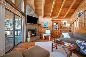 Ein rustikales Holzhaus-Interieur mit Kamin aus Stein, einem darüber montierten Fernseher, einem Sofa, einem Sessel und einer Terrasse mit Glastür.