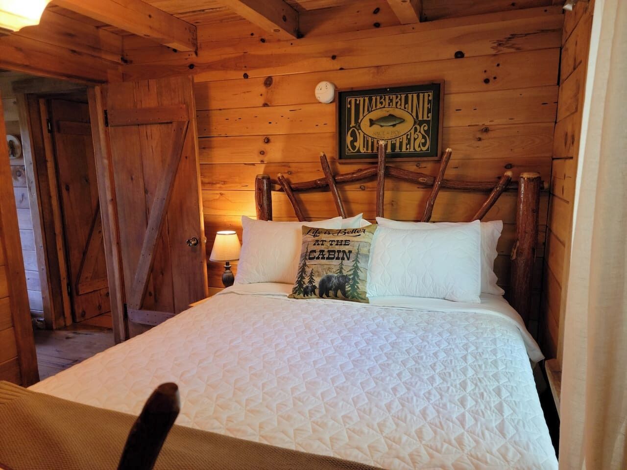 A wooden cabin-style bedroom with a bed, a pillow with a bear and forest design, and a sign above the bed reading 'Timberline Quarters.'