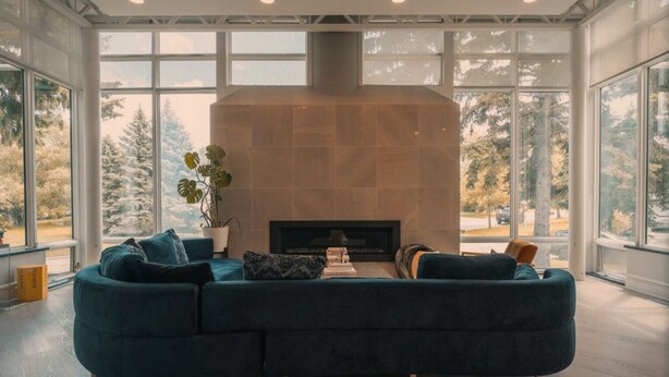Ein modernes Wohnzimmer mit einer großzügigen Eckcouch, einem Kamin und großen Fenstern mit Blick auf Bäume.