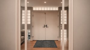Eine doppelte Tür mit Fußmatte davor, eine helle Wand und ein Fenster mit Jalousien.