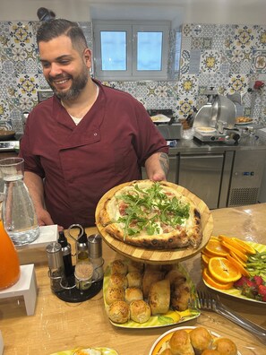 Una cucina con una pizza su un tagliere di legno, un piatto di panini e un vassoio di frutta.