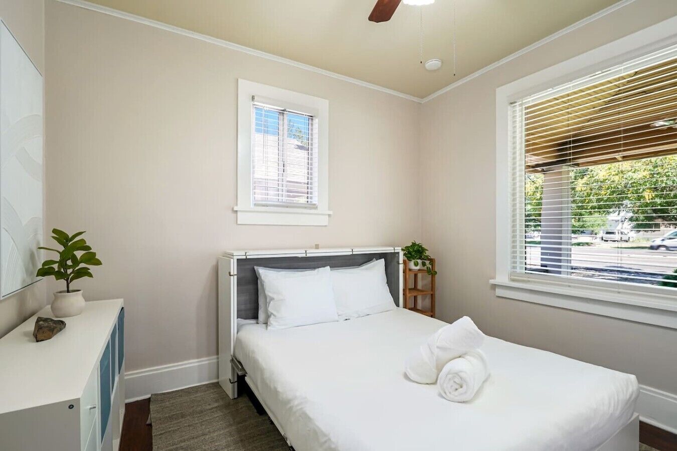 A bedroom with a bed, a bedside table with a plant, a window with blinds, and a ceiling fan.