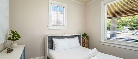 A bedroom with a bed, a bedside table with a plant, a window with blinds, and a ceiling fan.