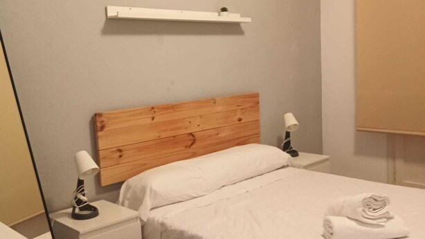 Un dormitori amb un capçal de llit de fusta, un llençol blanc, una taula de nit amb una làmpada i una petita estant sobre el llit.