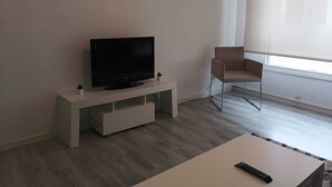 Un saló modern amb televisió de pantalla plana sobre un suport blanc, una cadira marró i una taula blanca amb un comandament a distància.