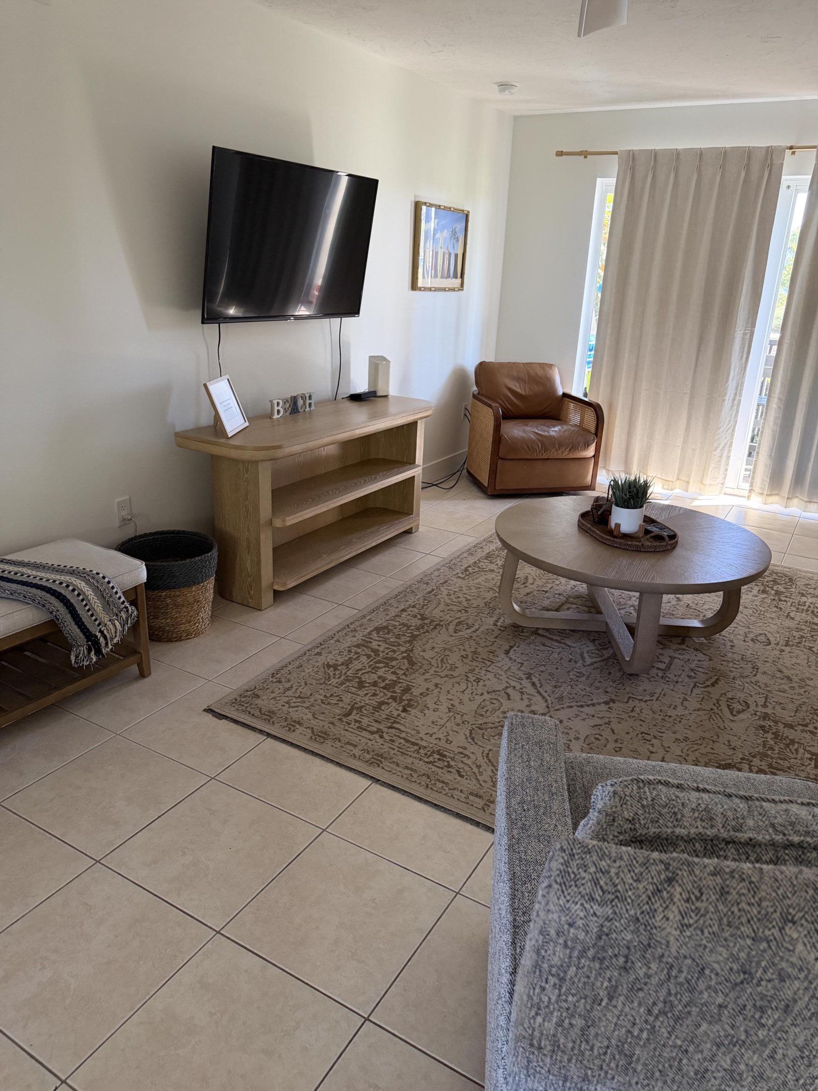 Een woonkamer met een flatscreen televisie, een houten tv-meubel, een leren fauteuil, een rond bijzettafeltje en een kamerbreed tapijt met een patroon.