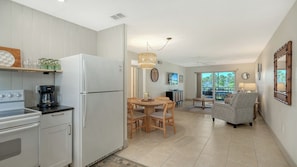 Eine moderne Küche mit weißen Geräten, ein Essbereich mit Holztisch und -stühlen sowie ein Balkon mit Blick auf Palmen.