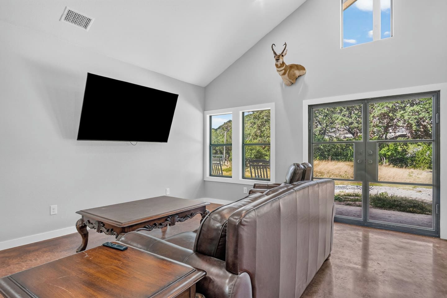 A modern living room with a flat-screen TV, a leather couch, a dining table, and a mounted deer head.