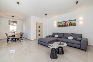 A modern living room with a grey sectional sofa, a dining table with chairs, a coffee table, and a painting of sailboats on the wall. - Villa ADRIA MOMENTS with private pool (Pinezići)