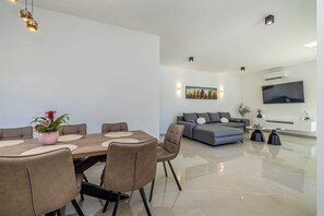 Ein modernes Wohnzimmer mit Sofa, Esstisch mit Stühlen, einem an der Wand montierten Fernseher und einem Gemälde über dem Sofa.