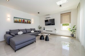 A modern living room with a grey sofa, a flat-screen TV, and a large window with blinds. - Villa ADRIA MOMENTS with private pool (Pinezići)