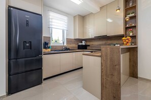 A modern kitchen with a black refrigerator, wooden cabinets, a built-in oven, and a window with blinds. - Villa ADRIA MOMENTS with private pool (Pinezići)