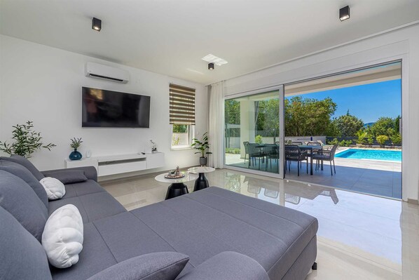 A modern living room with a large sofa, a flat-screen TV, and a glass door leading to an outdoor pool area.