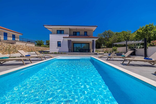 Ein moderner Poolbereich mit Liegestühlen, einem klaren, blauen Pool und einem zweigeschossigen Gebäude mit Naturstein-Fassade.