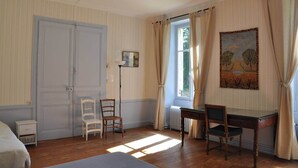 Una stanza con un letto, due poltrone, una scrivania, un termosifone, un quadro e una finestra con tende.