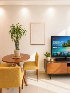 A modern living room with a dining area, a potted plant, a framed artwork, and a TV displaying a tropical paradise scene. - AÜRA - Beachfront Sųites + Pool @ Isla Verde (Carolina)