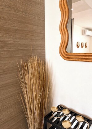 A room with a wooden textured wall, a tall decorative plant, a mirror with a wavy frame, and a black and white striped tray with bowls. - AÜRA - Beachfront Sųites + Pool @ Isla Verde (Carolina)