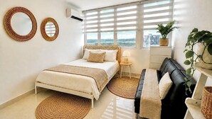 Ein Schlafzimmer mit einem Bett, einer schwarzen Couch, einem Fenster mit Jalousien und einer Zimmerpflanze.