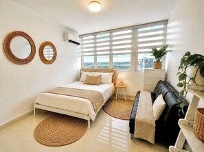 A bedroom with a bed, a black sofa, a window with blinds, and a potted plant. - AÜRA - Beachfront Sųites + Pool @ Isla Verde (Carolina)