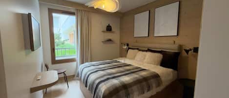 Un dormitorio con cama, escritorio, silla, ventana con cortinas y dos cuadros en la pared.