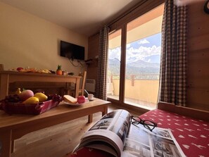 A room with a wooden table, a bench, a bed with a red bedspread, a window with a view of mountains, and a flat-screen TV mounted on the wall. - 50m from ski slopes, 2nd floor, swimming pool, balcony, ski locker, 32m², Vars (Vars)