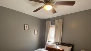Un dormitorio con cama, mesita de noche, ventilador de techo, ventana con cortinas e imagen colgada en la pared.