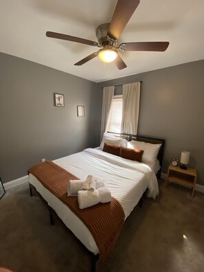 Ein Schlafzimmer mit einem Bett, einem Nachttisch, einem Deckenventilator, einem Fenster mit Vorhängen und einem Wandbild.