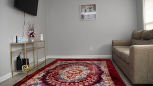 A living room with a sofa, a red patterned rug, a wall-mounted TV, and a small shelf with decorative items. - Charming 3-bedroom home with AC in Raleigh (Raleigh)