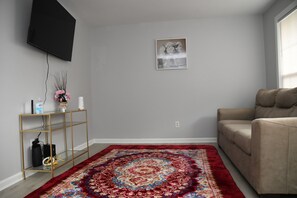 Una sala de estar con un sofá, un tapete rojo estampado, un televisor montado en la pared y una pequeña estantería con objetos decorativos.