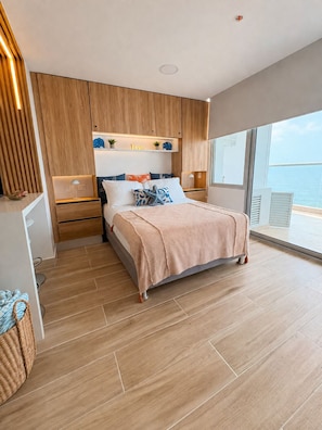 A modern bedroom with a large bed, wooden paneling, a nightstand, a bedside table, and a view of the sea. - Ocean View Suite in Cartagena 🌊 Pool • Beach • Airport in Crespo (Cartagena de Indias)