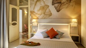 Ein Hotelzimmer mit einem großen Bett, zwei Nachttischlampen, einer gemusterten Bettdecke, Wandmalerei und einer Tür zu einem anderen Raum.