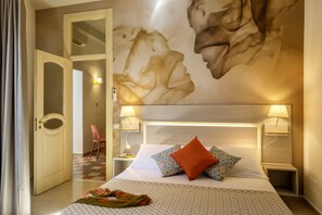 Ein Hotelzimmer mit einem großen Bett, zwei Nachttischlampen, einer gemusterten Bettdecke, Wandmalerei und einer Tür zu einem anderen Raum.