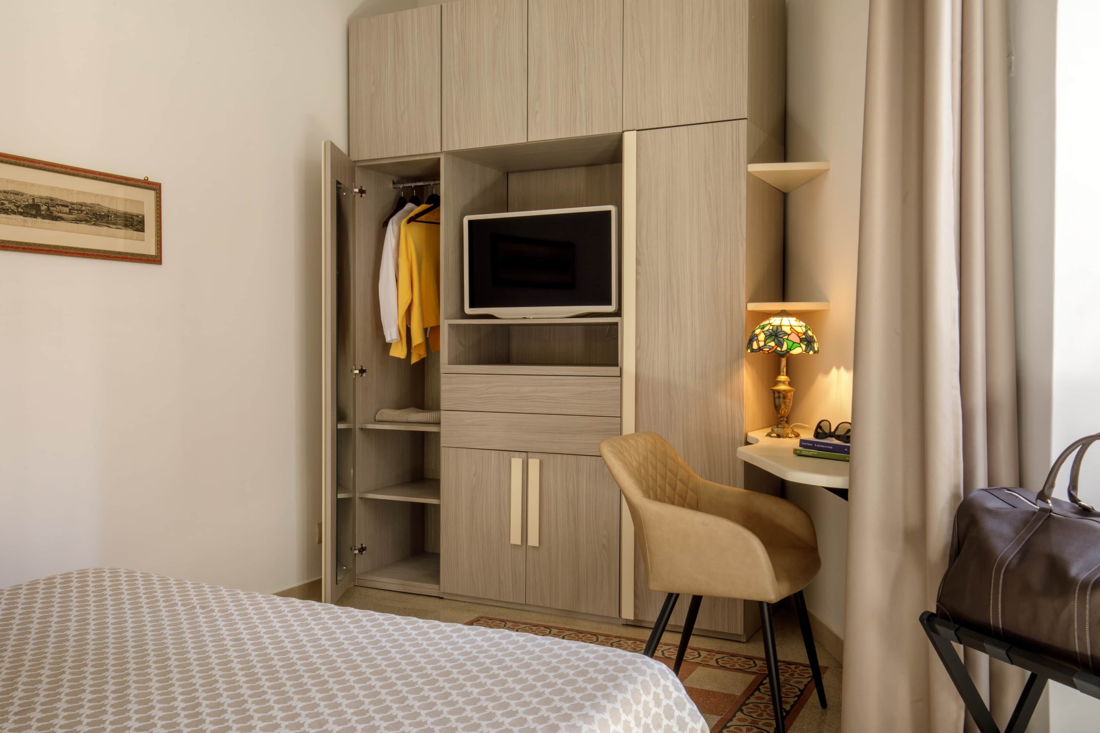A hotel room with a bed, a wardrobe with a TV, a desk with a lamp, and a chair.